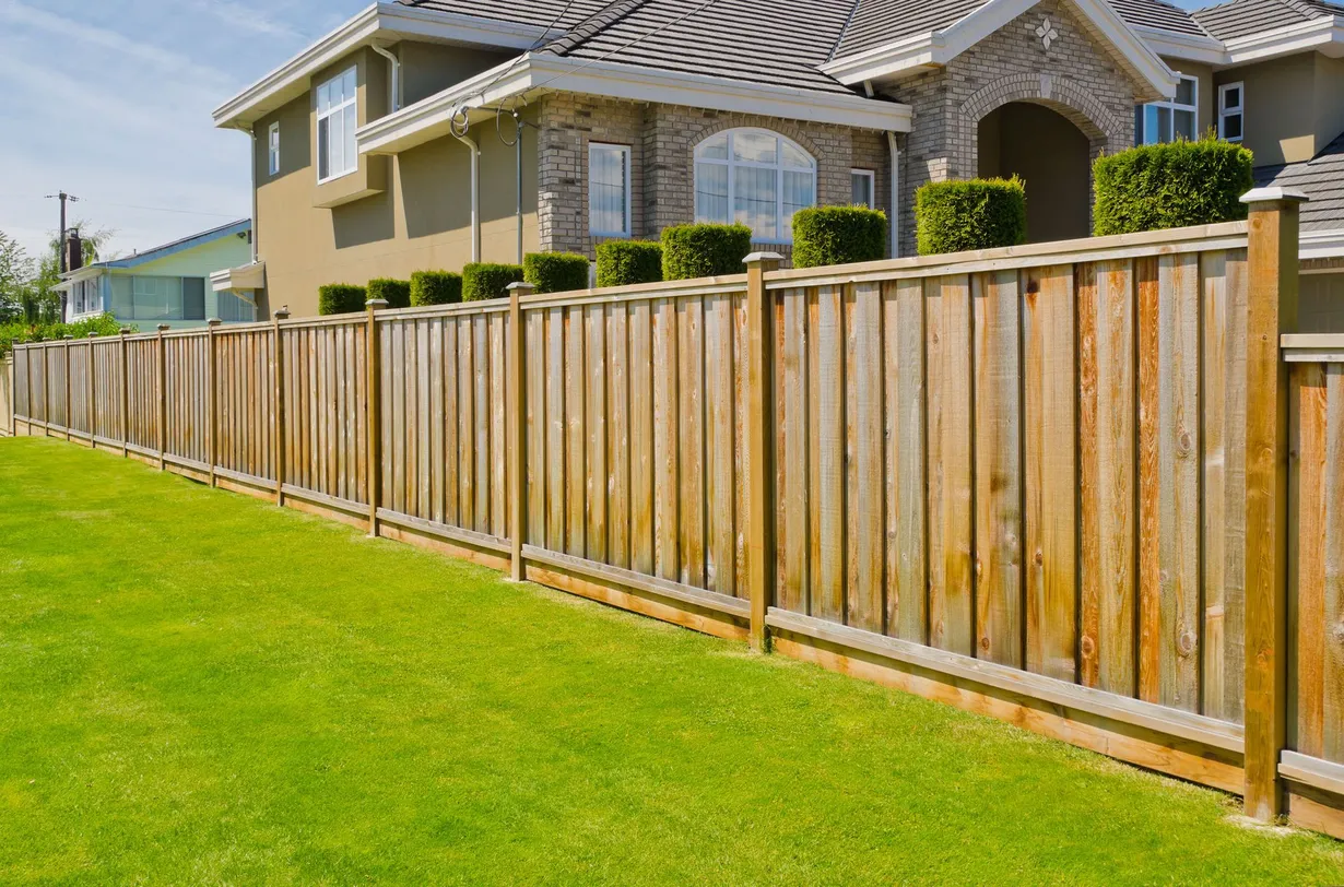 Wooden fence bordering a green lawn and a two-story house with beige siding.