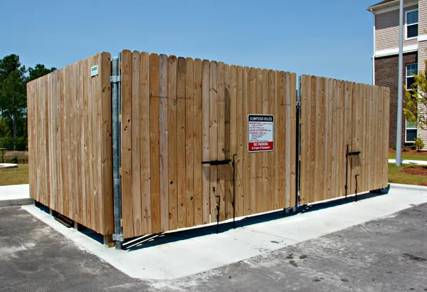 Wooden enclosure with gates containing dumpsters on a concrete pad. A warning sign is visible.