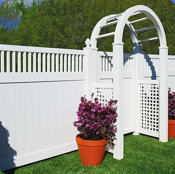 White picket fence with an arched gate, potted flowers. Green grass, blue sky.