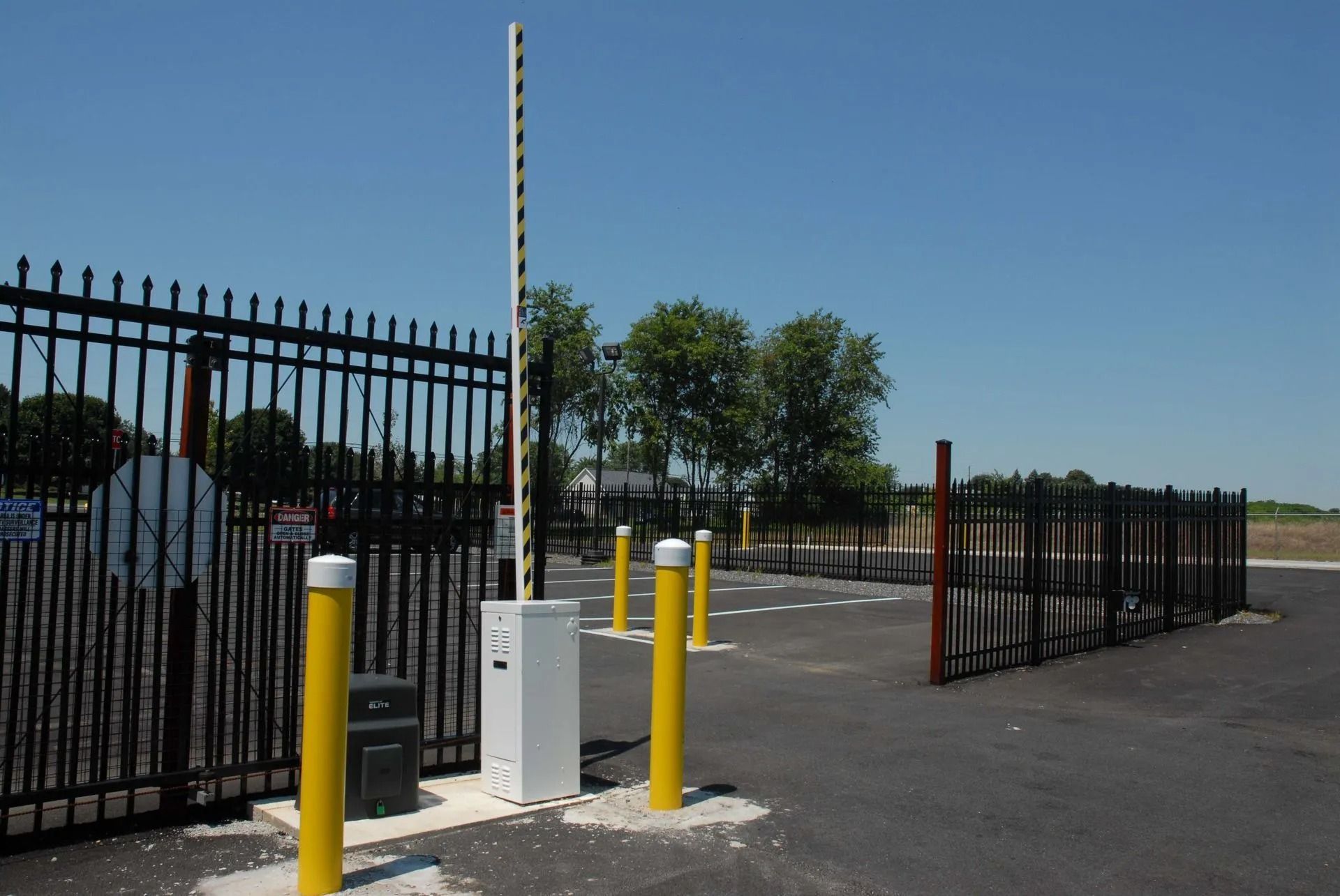 Black metal gate with raised arm, yellow bollards, and asphalt driveway under blue sky.