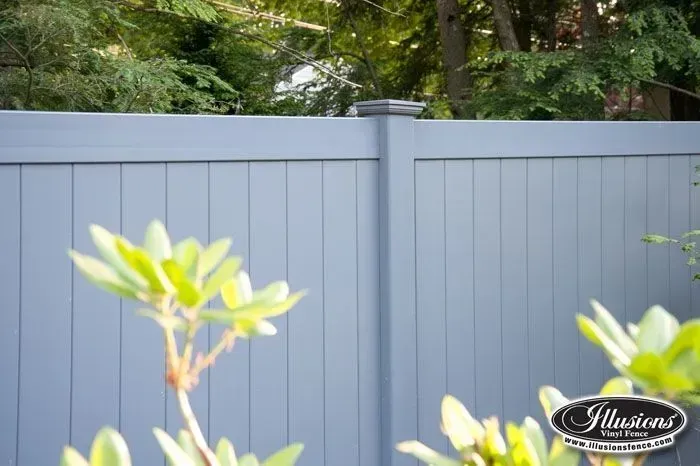Blue vinyl privacy fence with trees in the background, out of focus foreground with green plants.